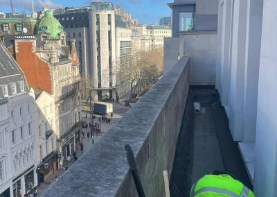 Worker installing floor screed onto a narrow ledge, high up over London, with view down onto a busy London street